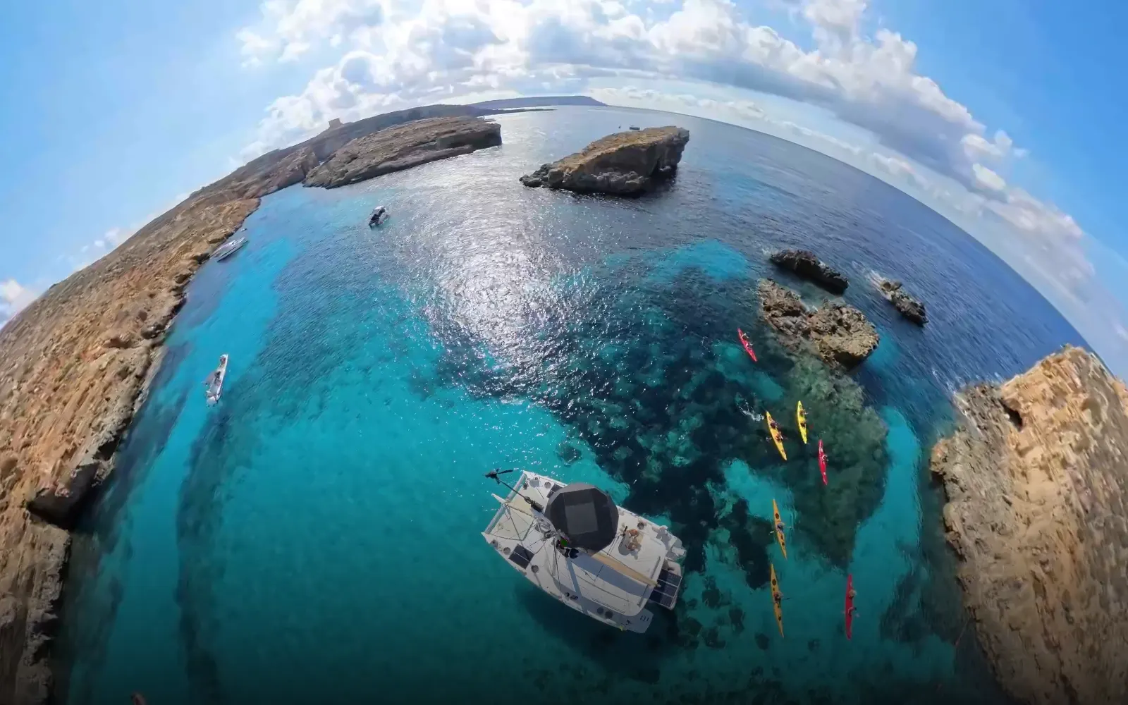 360° panorama over the Blue Lagoon, Comino — turquoise water, kayakers, a moored catamaran and limestone cliffs.